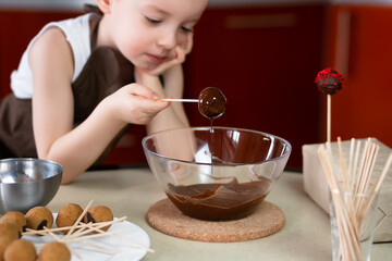 the child prepares cake pops at home in the kitchen, dips the billet in melted chocolate, the focus is on dessert - obrazy, fototapety, plakaty