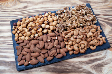 Various nuts sorted in glass bowl with honey bowl and honey dipper. Mixed nuts on wooden table. Black stone plate on wood background.