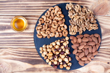 Various nuts sorted in glass bowl with honey bowl and honey dipper. Mixed nuts on wooden table. Black stone plate on wood background.