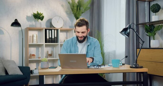 Handsome smiling confident skilled young bearded man in casual clothes working on laptop in beautifully decorated home office