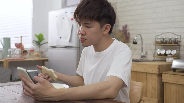asian boy sitting alone with a smartphone is phubbing while drinking tea and eating toast in the morning at home.