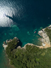 Aerial view of red boat sailing near steep stone cliff with green forest above transparent emerald water of Adriatic sea. Stones on seabed. Unspoiled nature of Montenegro coast. Budva scenic landscape