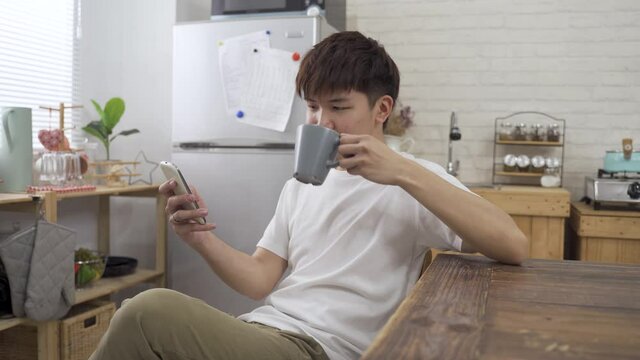 portrait japanese young guy is watching online videos on his smartphone while sitting at dining table and sipping coffee in leisure.