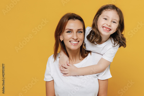 Happy woman in white t-shirt have fun with cute child baby girl 5-6 years old stand behind Mommy little kid daughter isolated on yellow orange color background studio Mother's Day love family concept