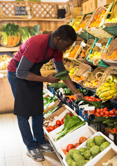 Experienced African American salesman filling supermarket counter with fresh vegetables and fruits..