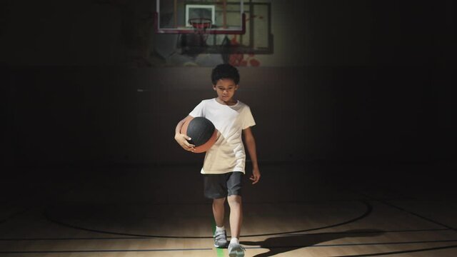 Slowmo portrait of little boy with ball walking up to camera and posing on dark basketball court with spotlight shining on him