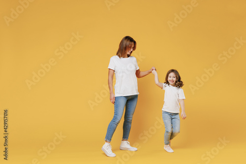 Full body length happy woman in basic white t-shirt have fun child baby girl 5-6 years old hold hands dance Mom little kid daughter isolated on yellow color background studio Mother's Day love family
