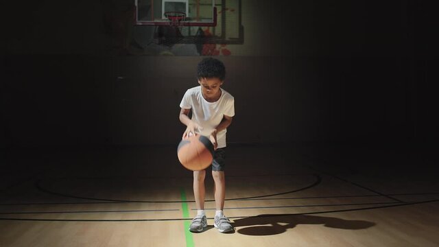 Slowmo shot of cute boy standing in spotlight on dark indoor basketball court and dribbling ball