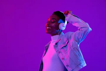 Zelfklevend Fotobehang Young African American woman wearing headphones listening to music and dancing in futuristic purple cyberpunk neon light background © Atstock Productions