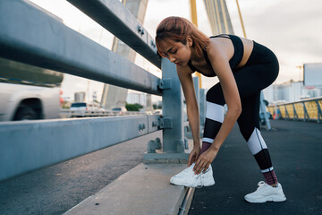 Young sporty fit Asian woman tying shoelaces of running shoes. Female runner working out at bridge and taking break between training in the fresh air