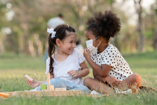 African little girl wear face mask talking to small Caucasian kid while sitting and playing wooden blocks toy in green park. Ethnic diversity of friendship in new normal lifestyle concept