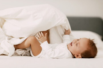 Fototapeta na wymiar The kid lies in a snow-white bed under the covers, laughs and indulges