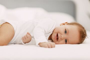 A blue-eyed baby lies in a snow-white bed under a blanket, laughs and indulges