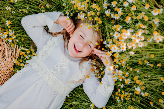 happy little girl in a cotton dress lies in a field of daisies in the summer at sunset. laughs, view from above