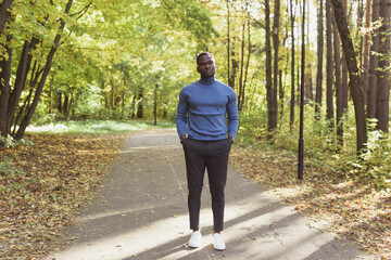 Fototapeta premium Positive african american young man in stylish clothes walks in late summer park on sunny warm warm day. Outdoor activity concept.