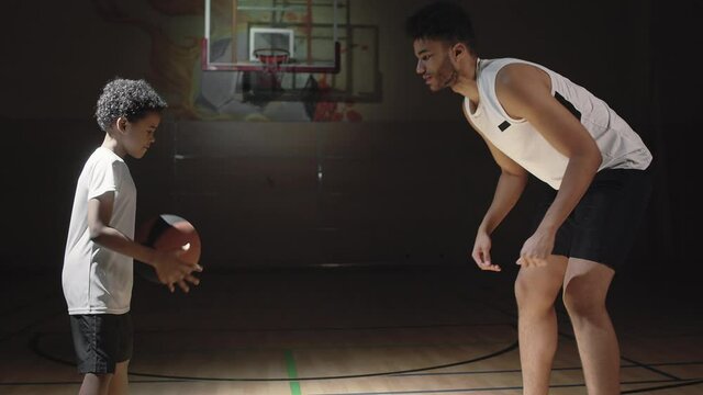 Medium shot of professional male basketball player teaching boy how to dribble on dark indoor court