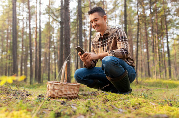 Naklejka na ściany i meble picking season and leisure people concept - happy smiling middle aged man with wicker basket and smartphone using app to identify mushroom in autumn forest