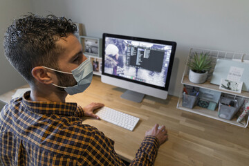 man works from home with his computer, dark-haired man in a mask and a plaid shirt writes on his home computer on a wooden desk