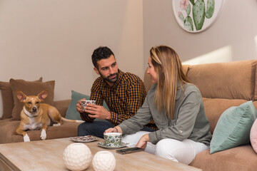 couple have a coffee, young couple sitting on a sofa having a coffee, next to it is lying their dog that is tan and white with short hair