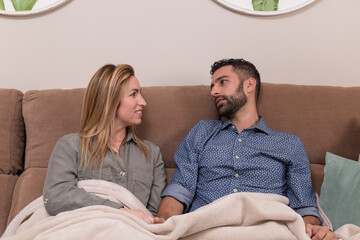 couple sitting on the sofa and covered with a blanket look at each other with complicity