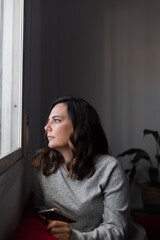 woman looking out the window, pensive woman observes the street through a closed window she has a mobile in her hand, vertical photo