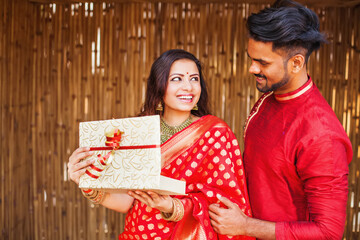Indian man in festive ethnic clothes giving a wrapped gift box to a woman in a red saree, she is smiling and feeling happy