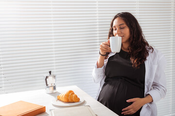 Happy pregnant woman eating croissant during morning breakfast. Concept of pleasant morning and positive attitude during pregnancy