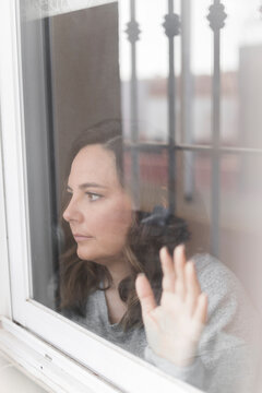 woman looking out the window, pensive woman observes the street through a closed window and leaning her hand on the glass, the bars of the house are reflected in the glass, vertical photo