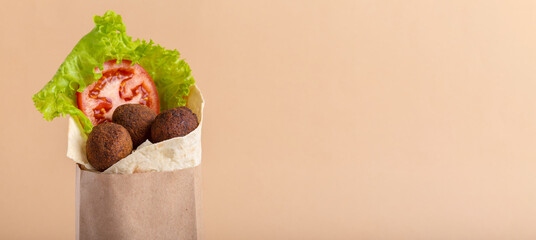 Horizontal banner with tasty falafel pita roll with ripe red tomato and green salad in craft paper bag for delivery on beige background. Copy space. Healthy vegetarian fastfood
