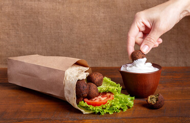 Woman hand dipping fried falafel into yogurt sauce in clay bowl on brown board with burlap background. Falafel pita roll with ripe red tomato and green salad in craft paper bag. Jewish cuisine