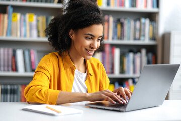 Satisfied african american smart female student or freelancer, with curly hair, trendy dressed, is sitting at a work desk, uses a laptop, concentrated browsing internet, searching information, smiling