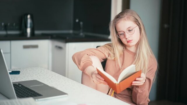 Beautiful 10 years old blond girl in glasses sitting at the desk and reading a book while doing her homework.