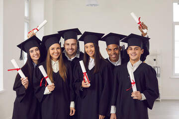 Graduation from university concept. Group of happy multiracial students university graduates standing and looking at camera with dimplomas in hands over white wall in class at background
