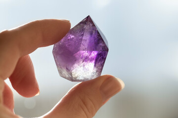 The amethyst crystal is held by the fingers of the hand. Purple semi-transparent faceted crystal close-up