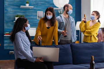 Multi ethnic friends wearing face mask drinking beer using laptop keeping social distancing to prevent coronavirus spread during global pandemic having fun in home living room. Conceptual image.