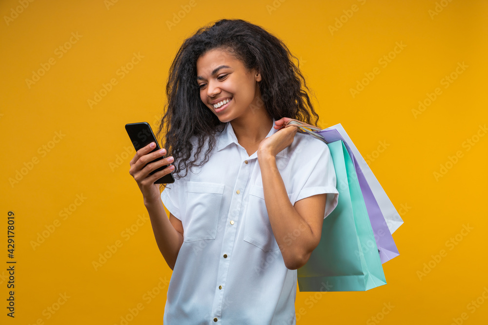 Wall mural portrait of beautiful young dark skinned woman with shopping bags using her smart phone on bright ye