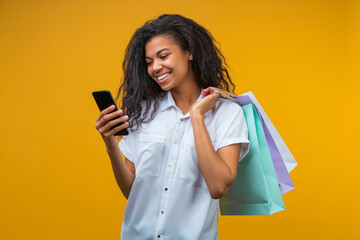 Portrait of beautiful young dark skinned woman with shopping bags using her smart phone on bright yellow background