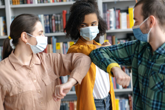 Happy student friends wearing protective face masks greeting bumping elbows while standing at university library, healthcare concept, precautions, new normal between people communication