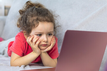 A pretty little curly girl is lying in front of a laptop and watching a cartoon. Rest at home, child and computer.