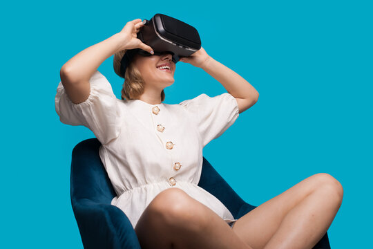 Caucasian woman sitting on armchair and testing new vr headset smiling on a blue studio wall