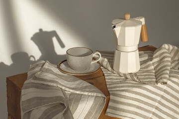 Italian coffee maker with wooden handle and white coffee craft cup on solid wood stamp decorated with tea towels in creamy tones on sunlight creating beautiful shadows on the wall