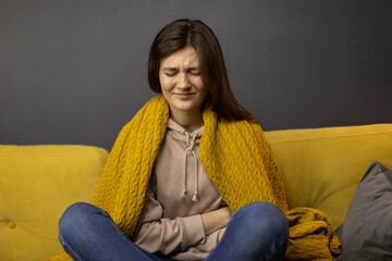 Attractive long-haired brunette girl sits on yellow sofa knees bent suffering from stomach ache at...