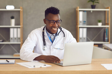 Happy medical specialist working on computer. Handsome black doctor sitting at office desk and...