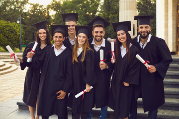 Group of graduate students in robes and mortarboards holding their diplomas near university campus....