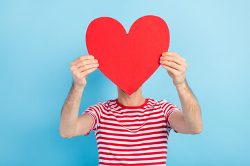 Photo of sweet shy young man dressed striped t-shirt red paper heart cover face head empty space isolated blue color background