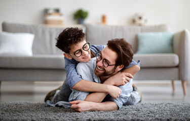 Father son bonding. Daddy lying on carpet carrying boy on back and smiling, having fun at home