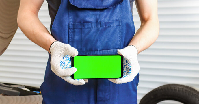 A Man In A Blue Jumpsuit Holds A Phone With A Green Screen In A Horizontal Position. Auto Mechanic In Gloves In The Garage Near The Car Repair.