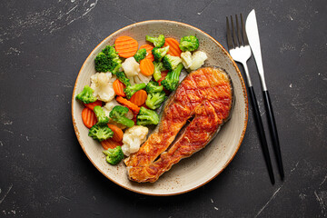 Healthy fish dinner: grilled salmon fish steak with vegetables salad on ceramic plate with fork and knife on black slate stone background from above, healthy clean and diet eating concept 