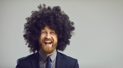 Obraz premium Copy space banner with closeup portrait of excited young man in office suit and funny wig looking at camera and laughing. Headshot of happy guy with crazy curly hair isolated on gray studio background