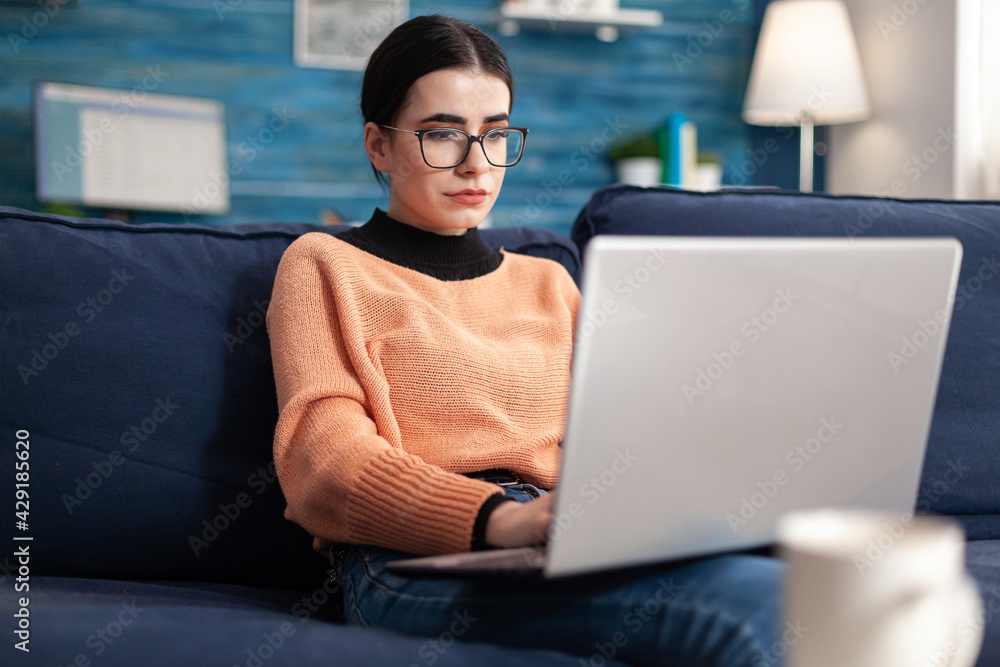 Poster college student with glasses checking email on laptop computer while sitting on sofa in living room.